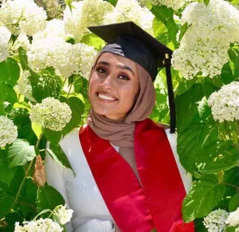 Smiling woman in beige hijab, black cap, white graduation gown, and red stole