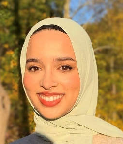Photo of smiling woman with light colored hijab with fall foliage in the background