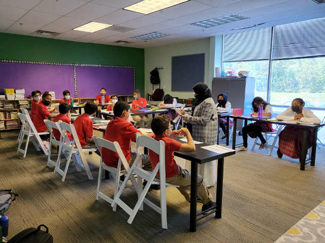 [Image Description] Classroom with students sitting around the table while the teacher leads instruction between the tables.