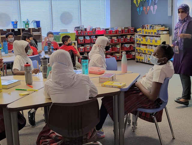 [Image Description: Teacher in lavender clothing and scarf teaches students sitting at a table in the classroom.
