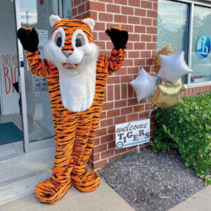 Photo of school mascot tiger standing in front of building with sign in ground that says Welcome, TIgers